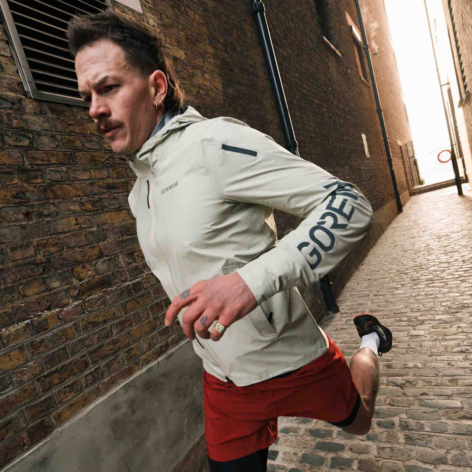 A model running in the GOREWEAR Men's Concurve GORE-TEX Logo Jacket in the Tech Beige/Black colourway. (8751737962658)
