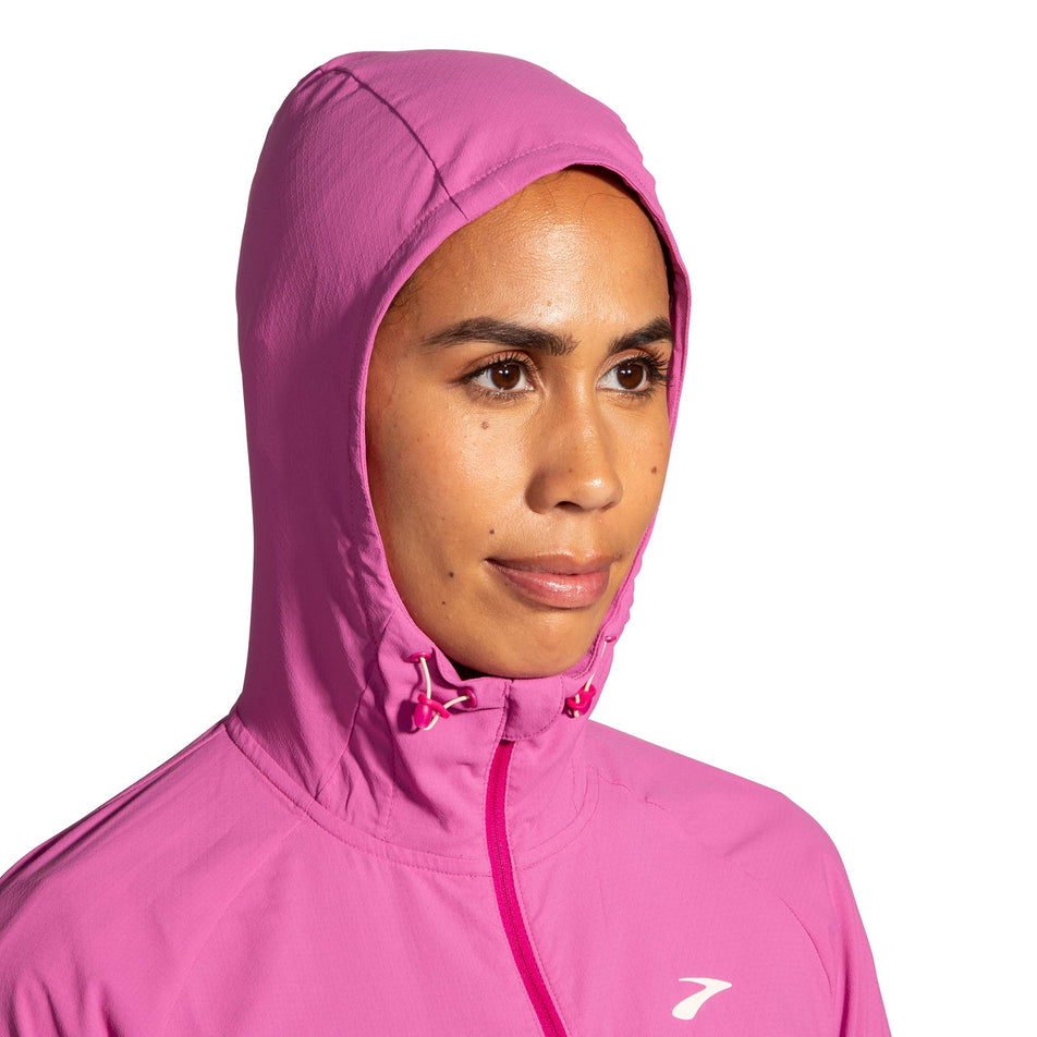 Close-up front view of a model wearing a Brooks Women's Canopy Jacket in the Frosted Mauve/Mauve colourway. Upper third of the jacket is visible. Model is wearing the jacket with the hood up.  (8007491748002)