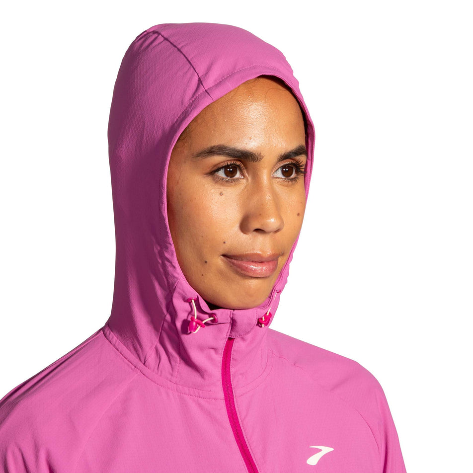 Close-up front view of a model wearing a Brooks Women's Canopy Jacket in the Frosted Mauve/Mauve colourway. Upper third of the jacket is visible. Model is wearing the jacket with the hood up. (8007491748002)