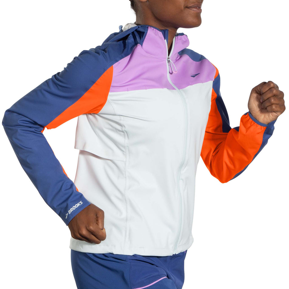 Right-side view of a model wearing a Brooks Women's High Point Waterproof Jacket in the Lt Slate/Bright Orange/Aegean colourway. Model is in a running pose. (8037744771234)