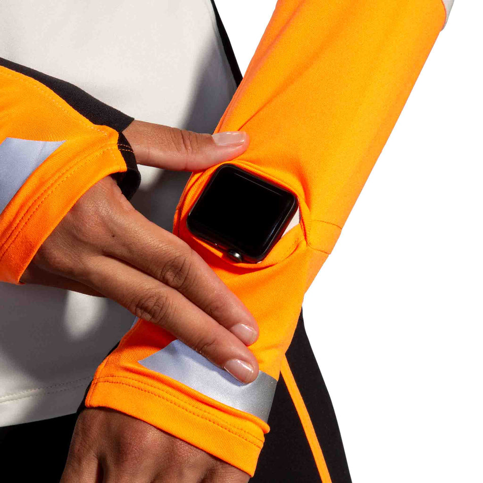 The watch window on the left sleeve of the Brooks Women's Run Visible 1/2 Zip 2.0 in the Ecru/Fluoro Flash/Black colourway. (8499447333026)