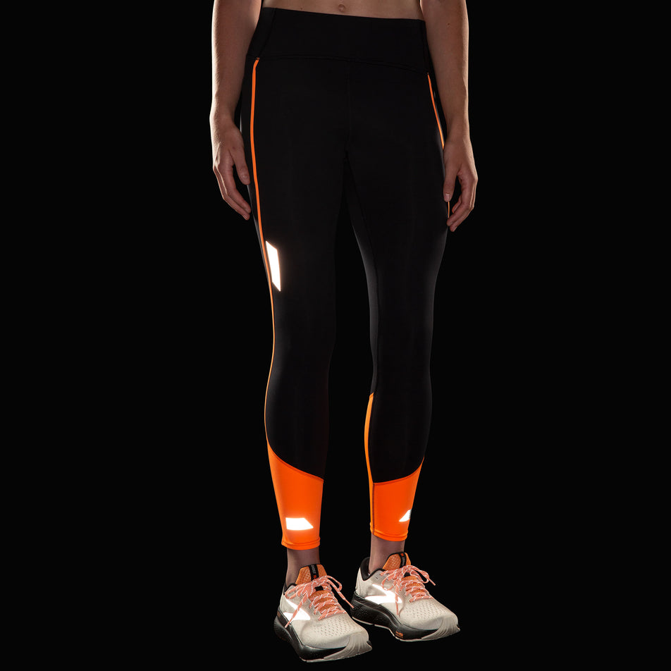 Front view of a model wearing the Brooks Women's Run Visible Tight 2.0 in the Black/Fluoro Flash colourway. Reflective strips are reflecting artificial light. Model is also wearing Brooks shoes. (8499453362338)