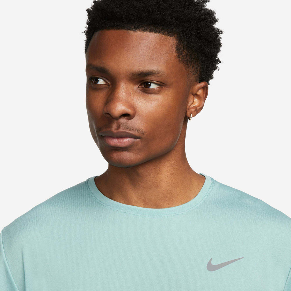Close-up front view of a model wearing a Nike Men's Miler Dri-FIT UV Short-Sleeve Running Top in the Mineral/Jade Ice/HTR/Reflective Silv colourway. T-shirt's neck, shoulder and chest sections are visible. (7980005261474)
