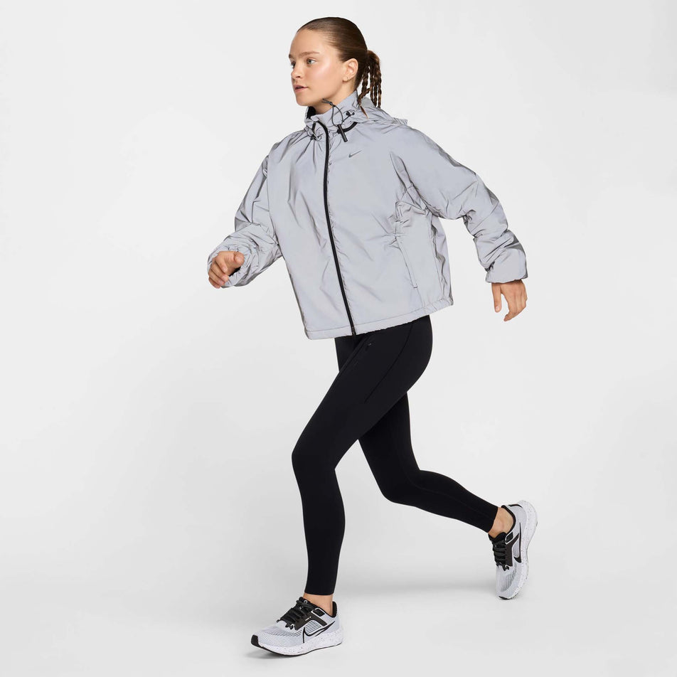 Angled view of the front of a model wearing Nike Women's Running Division Therma-FIT Reflective Running Jacket in the Black/Reflective Silv/Refblk colourway. Model is also wearing black Nike tights and Nike shoes. (8497648730274)