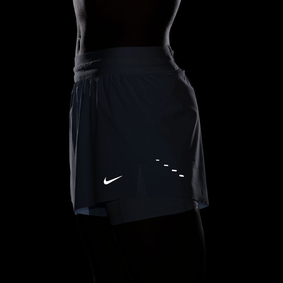 Side view of a model wearing a pair of Nike Women's Swift Dri-FIT Mid Rise 2-in-1 Running Shorts in the Blue Tint/Reflective Silv colourway. Photo has been taken in low light conditions to show the reflective details on the shorts. (8620122800290)