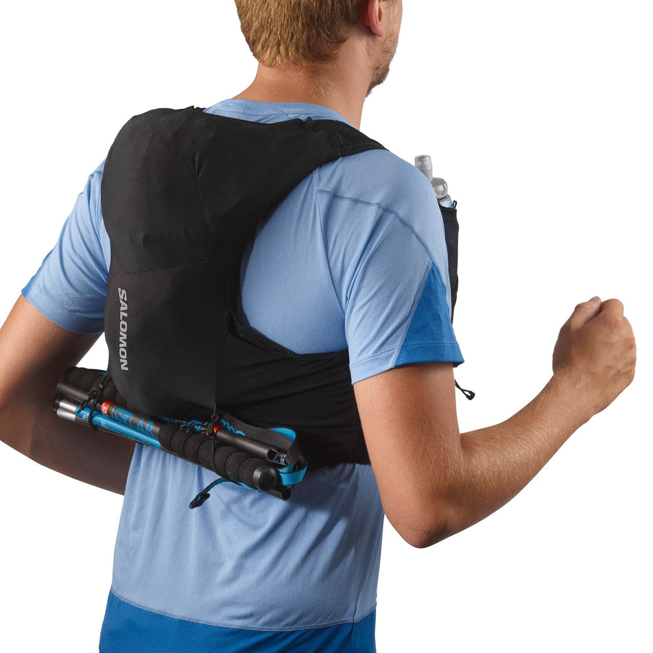 Back view of a model wearing a Salomon Unisex ADV Skin 5 Running Vest in the Black/Ebony colourway, with flasks included. Example of running poles being stored at the base of the back of the pack.  (7991863115938)