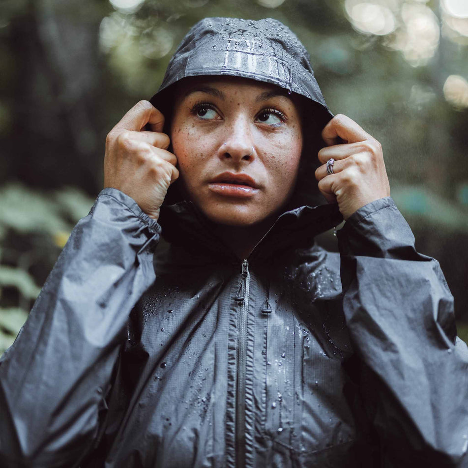 A model wearing a Salomon Bonatti Waterproof Shell Jacket in the Deep Black colourway. Model is wearing the jacket with the hood up, and there are beads of water on the jacket. (8464141320354)