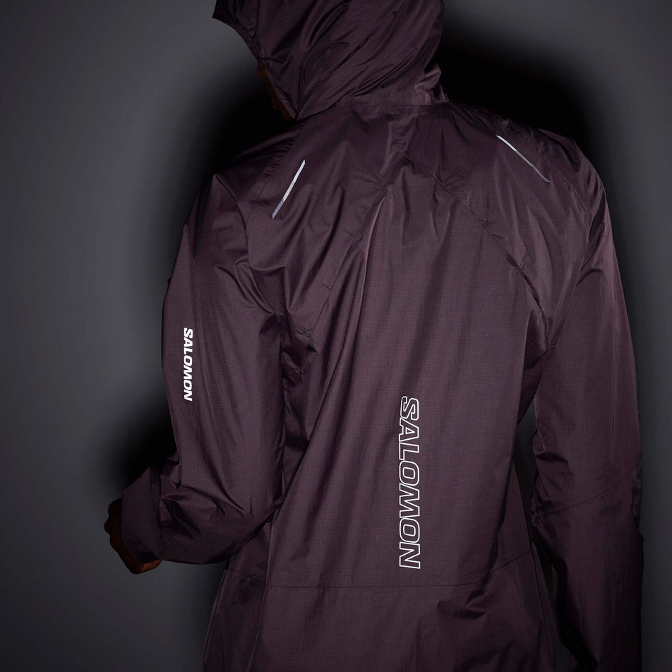 Back view of a model wearing a Salomon Women's Bonatti Waterproof Jacket in the Moonscape colourway, with the hood up. Being worn in low light conditions to demonstrate the reflective Salomon logo and reflective strips on the back of the jacket, and the reflective Salomon logo on the left sleeve.  (7999026266274)