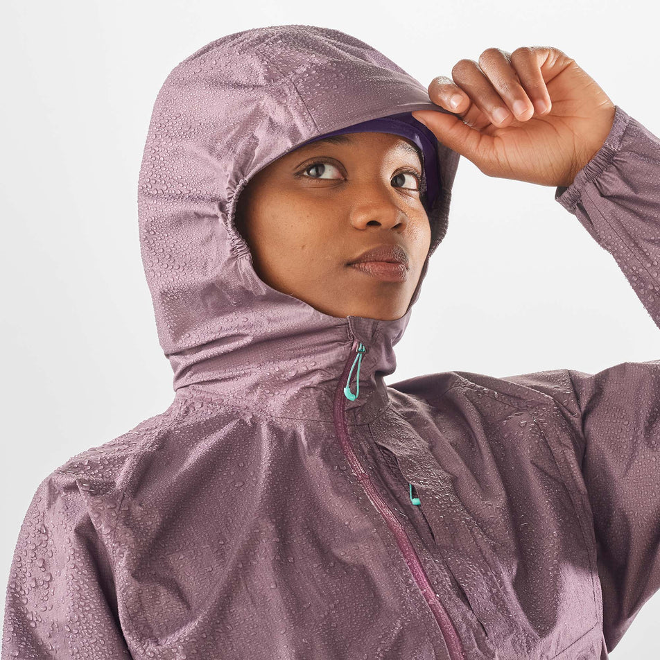 Close-up front view of a model wearing a Salomon Women's Bonatti Waterproof Jacket with the hood up. Upper third of the jacket is visible. Beads of water are sitting on the jackets surface. Jacket is in the Moonscape colourway.  (7999026266274)