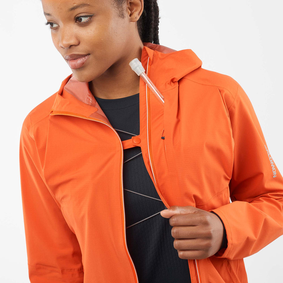 Close-up front view of a model wearing a Salomon Women's Bonatti Trail Jacket in the Burnt Ochre colourway. Jacket is unzipped, but the front sides are fastened together with the built-in clip.  (8000727351458)