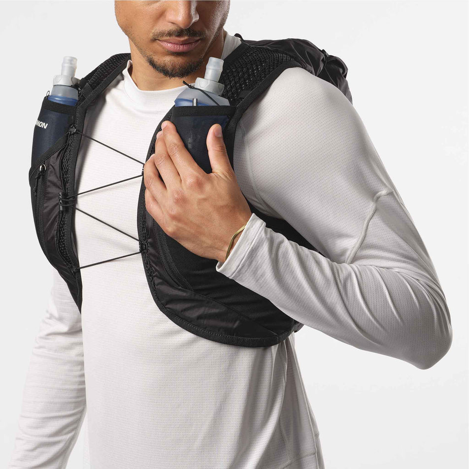 Front view of a model wearing a Salomon Unisex Active Skin 12 Set in the Black/Metal colourway. Model is highlighting the ease of access to a soft flask on the front of the vest. (8407345889442)