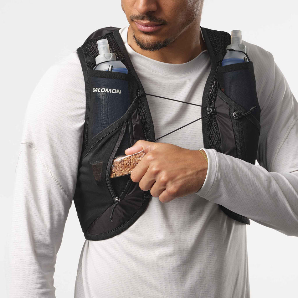Front view of a model wearing a Salomon Unisex Active Skin 12 Set in the Black/Metal colourway. Model is demonstrating the ability to store nutrition in one of the zipped pockets on the front of the vest. (8407345889442)