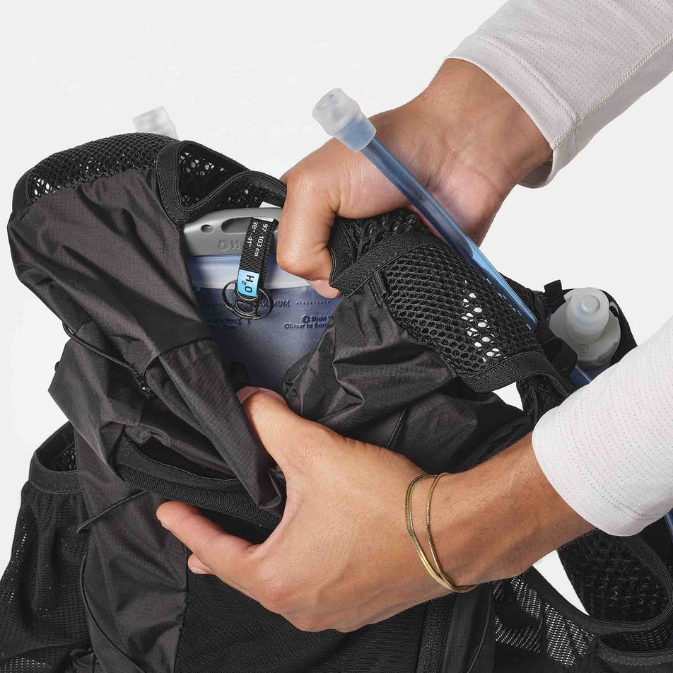 Model is demonstrating the ability to store a hydration bladder in the back of a Salomon Unisex Active Skin 12 Set in the Black/Metal colourway. (8407345889442)