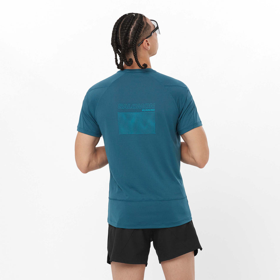 Back view of a model wearing a Salomon Men's Cross Run GFX Short Sleeve T-Shirt in the Deep Dive colourway. Model is also wearing shorts and sunglasses. (8157850534050)