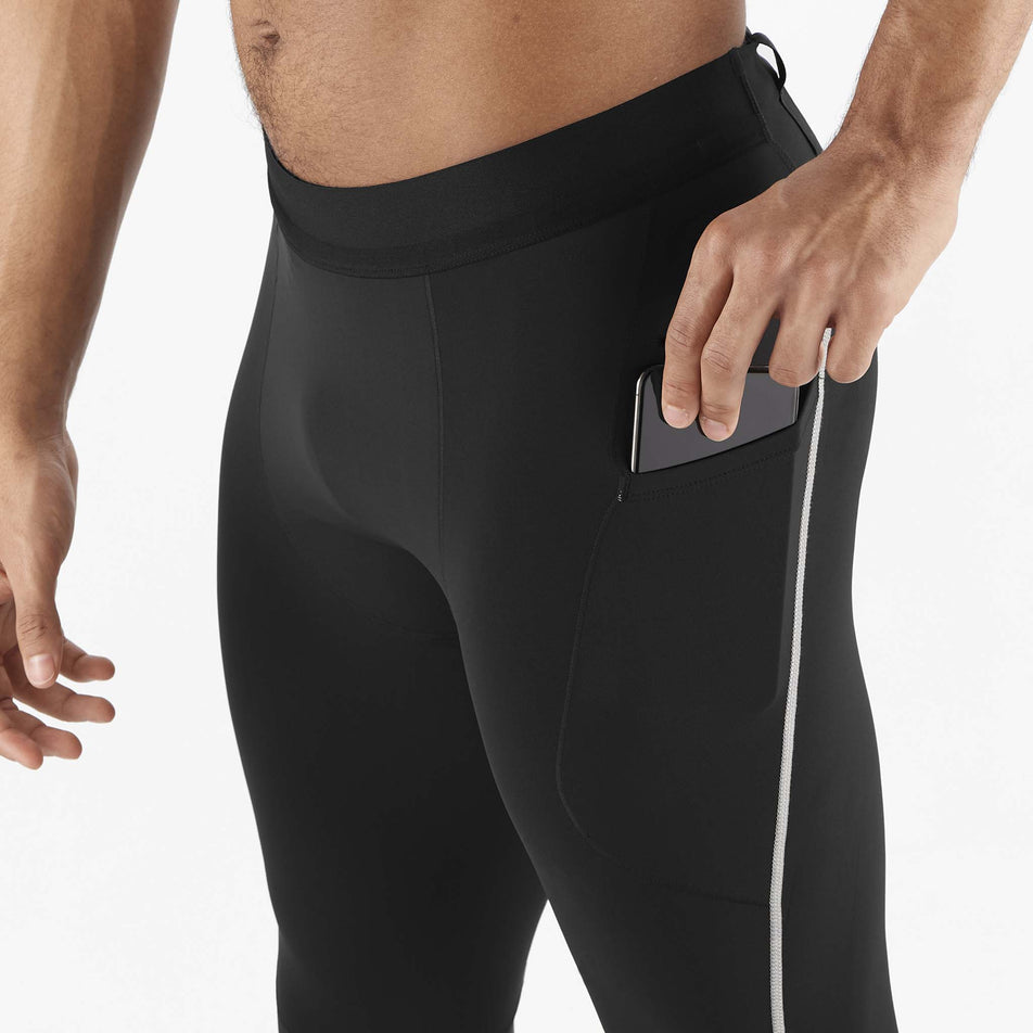 A model demonstrating that a phone can be stored in the stretch side pocket on the outside of the left-leg of a pair of Salomon Men's Sense Aero Stow Tights. Tights are being worn by a model. (8466531057826)
