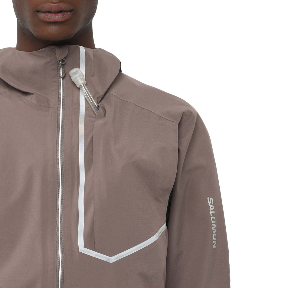 A model demonstrating the ability to have the drinking valve of a soft flask coming out the chest pocket of the Salomon Men's Bonatti Trail Shell Jacket in the Iron colourway. (8736773374114)