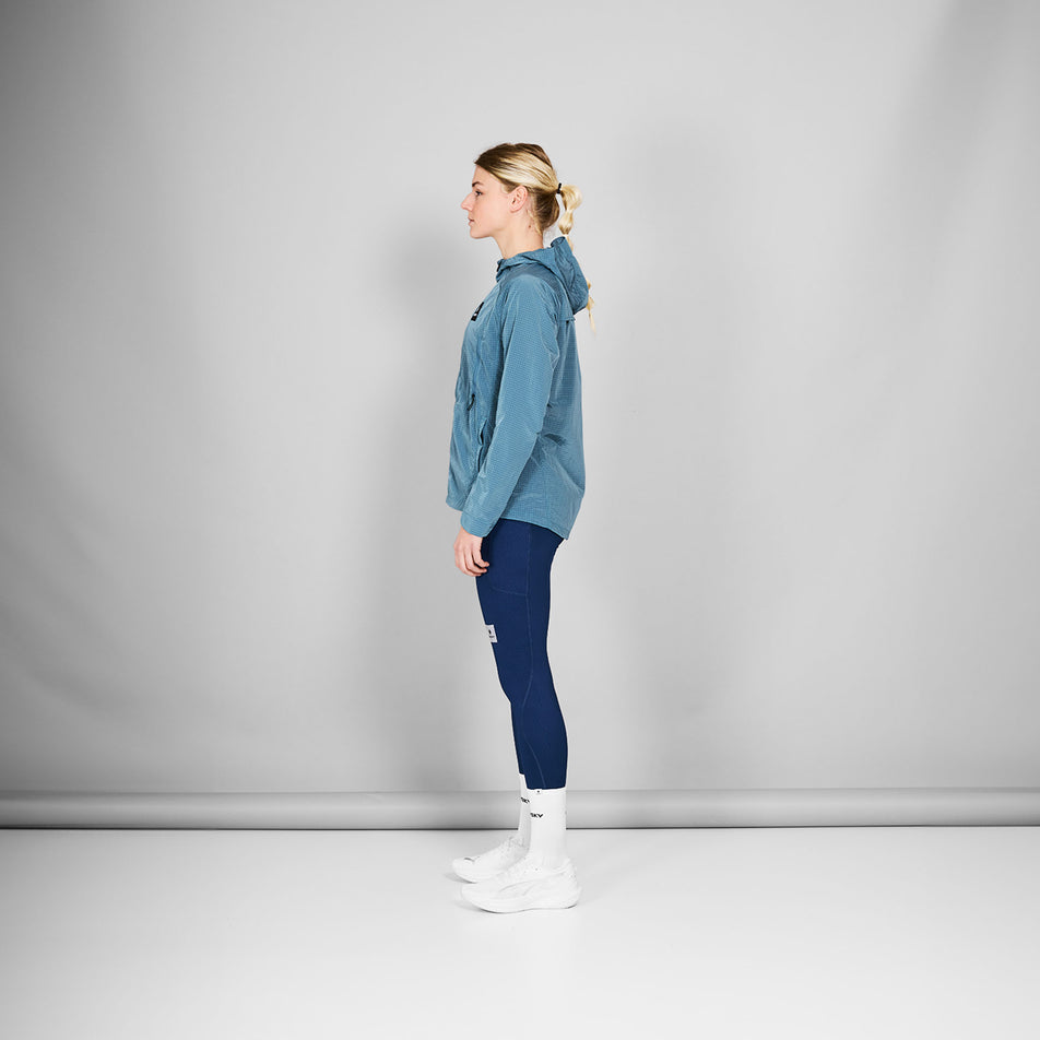Side view of a model wearing the SAYSKY Women's Clean Pace Shield Jacket in the Blue colourway. Model is also wearing tights, socks and shoes. (8601993445538)