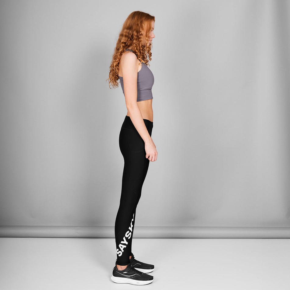 Side view of a model wearing the SAYSKY Women's Logo Motion+ Long Tights in the Black colourway. Model is also wearing shoes and a top. (8602027950242)