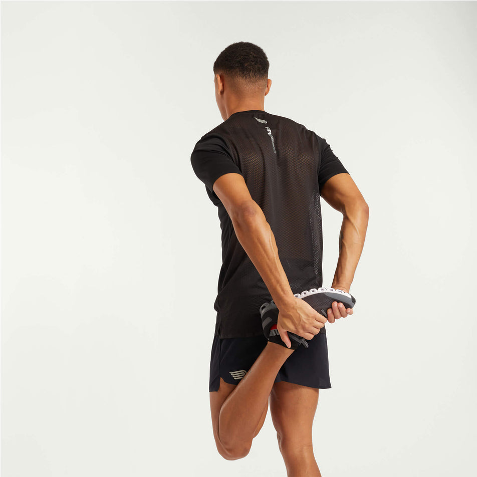 Back view of a model wearing a Pressio Men's Elite S/S Top in the black colourway. Model is also wearing running shoes, socks and Pressio shorts, and is in a stretching pose. (8248497373346)