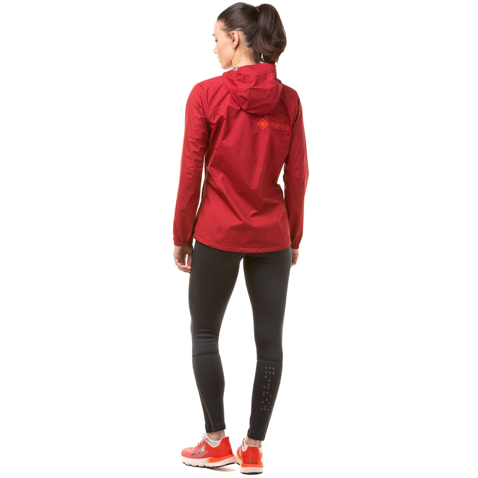 Back view of a model wearing a Ronhill Women's Tech GORE-TEX Mercurial Jacket in the Jame/Flame colourway. Model is also wearing black Ronhill running leggings and red Altra running shoes. (8047328034978)