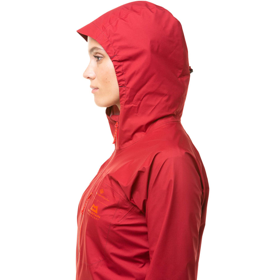 Close-up left-side view of a model wearing a Ronhill Women's Tech GORE-TEX Mercurial Jacket in the Jam/Flame colourway. Upper two-thirds of the jacket is in the image. Jacket is being worn with the hood up. (8047328034978)