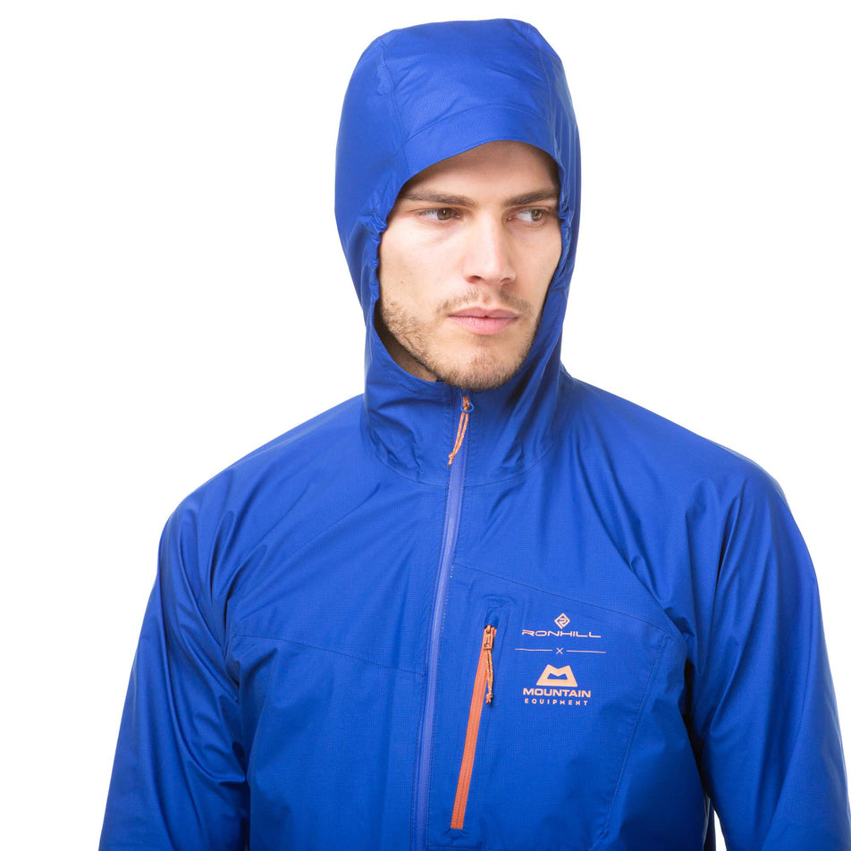 Close-up front view of a model wearing a Ronhill Men's Tech Gore-Tex Mercurial Jacket in the Cobalt/Copper colourway. Model is wearing the jacket with the hood up. Upper half of jacket can be seen in the image.  (8032222019746)