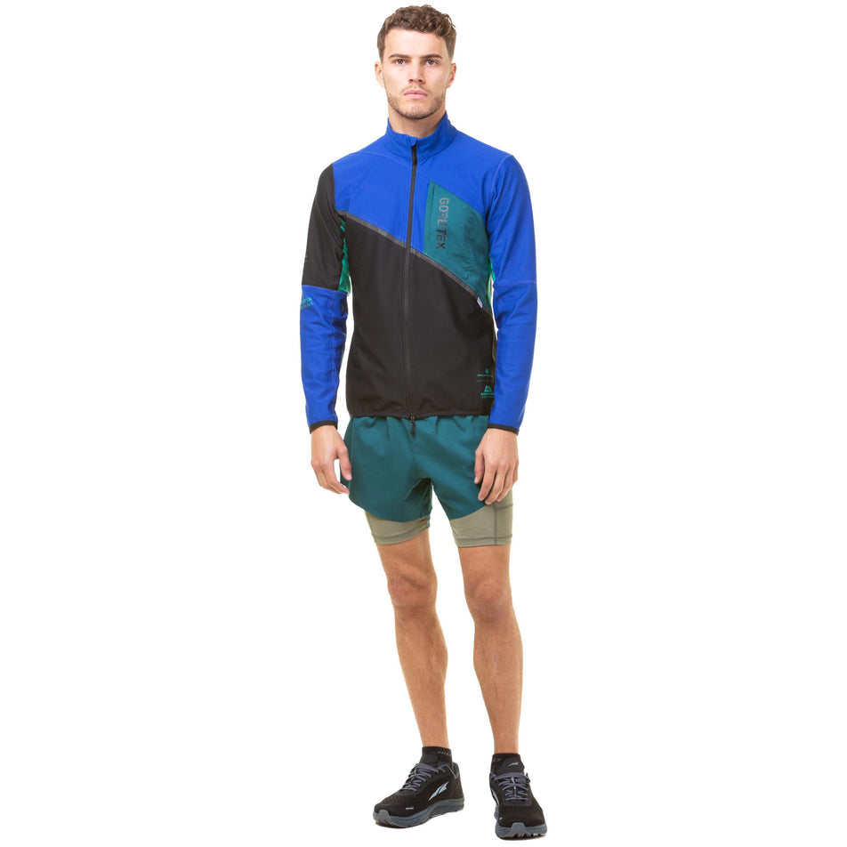 Front view of a model wearing a Ronhill Men's Tech Gore-Tex Windstopper Jacket in the Black/Cobalt colourway. Model also wearing a pair of green Ronhill shorts and black Altra running shoes.  (8032213565602)