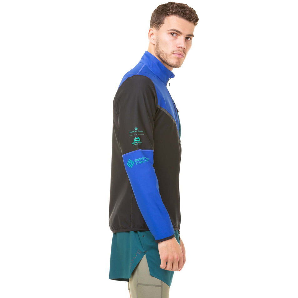 Side view of a model wearing a Ronhill Men's Tech Gore-Tex Windstopper Jacket in the Black/Cobalt colourway. Model also wearing a pair of green Ronhill shorts. (8032213565602)