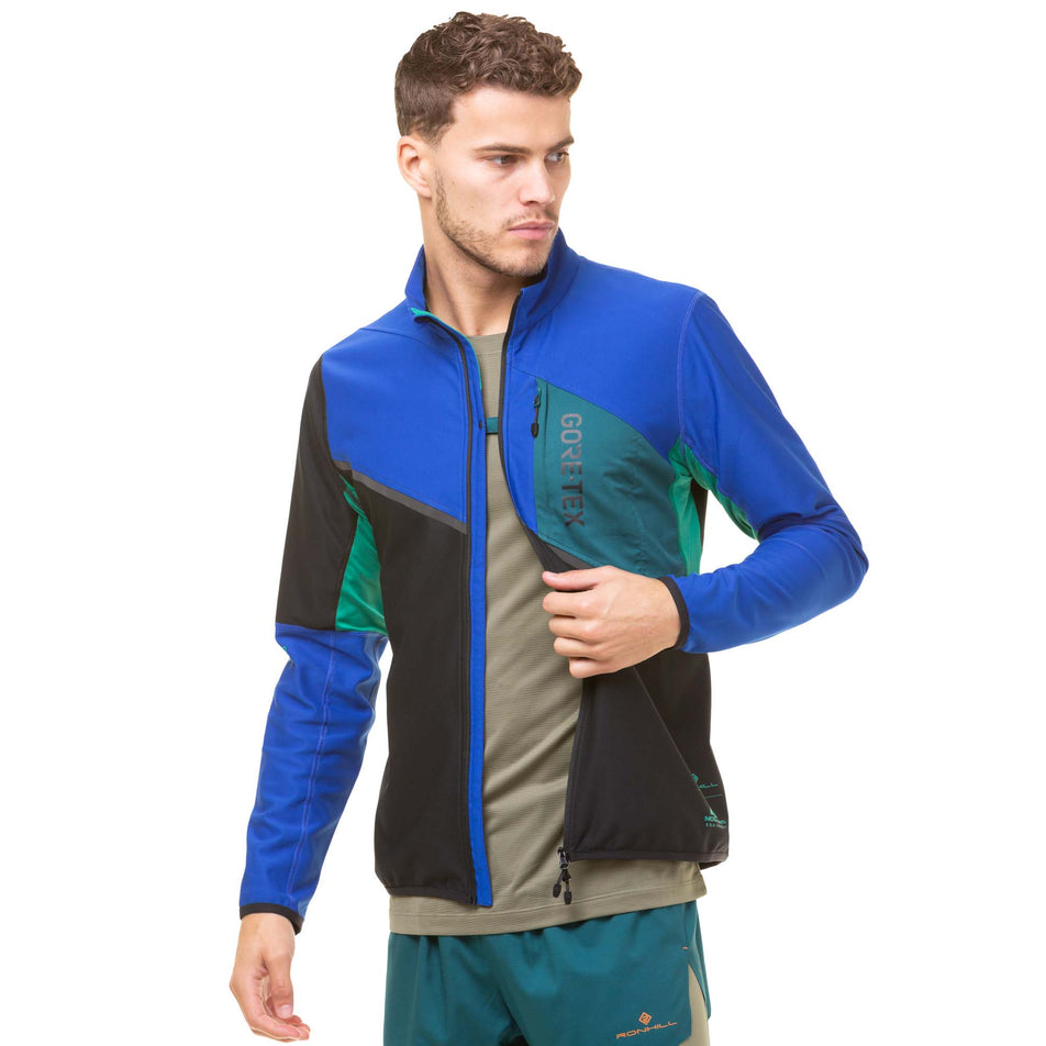 Front view of a model wearing a Ronhill Men's Tech Gore-Tex Windstopper Jacket in the Black/Cobalt colourway. Model is demonstrating that the front left and right sides of the jacket can be fastened together at the chest - whilst the jacket is unzipped. Model also wearing a green Ronhill t-shirt and pair of shorts.  (8032213565602)