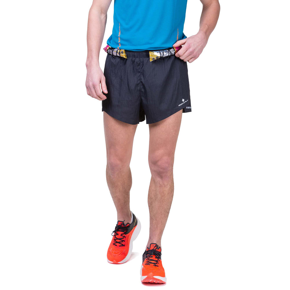 Front view of a model wearing the Ronhill Men's Tech Race Short in All Black colourway, and demonstrating that energy gels can be stored in the elasticated front loops. Model also wearing a Ronhill top and Altra running shoes.  (8160873119906)