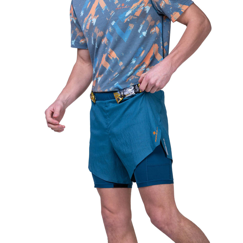 Front view of a model wearing the Ronhill Men's Tech Race Twin Short in the Legion/Fluo Orange colourway, and demonstrating that energy gels can be stored in the elasticated front loops. Model is also wearing a Ronhill top. (8160877543586)