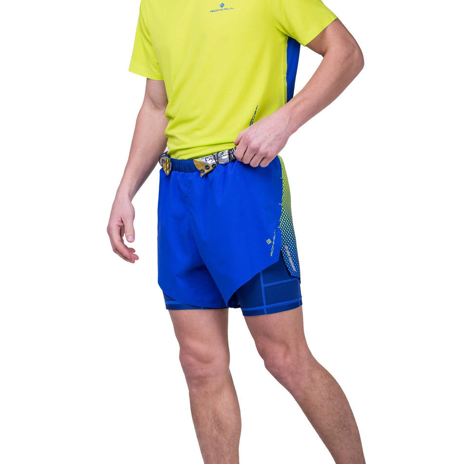 Front view of a model wearing the Ronhill Men's Tech Race Twin Short in the Azurite/Citrus Fade colourway. Model is wearing the shorts with energy gels in the elasticated loops on the front of the waistband, and is wearing a Ronhill running t-shirt too. (8270451409058)