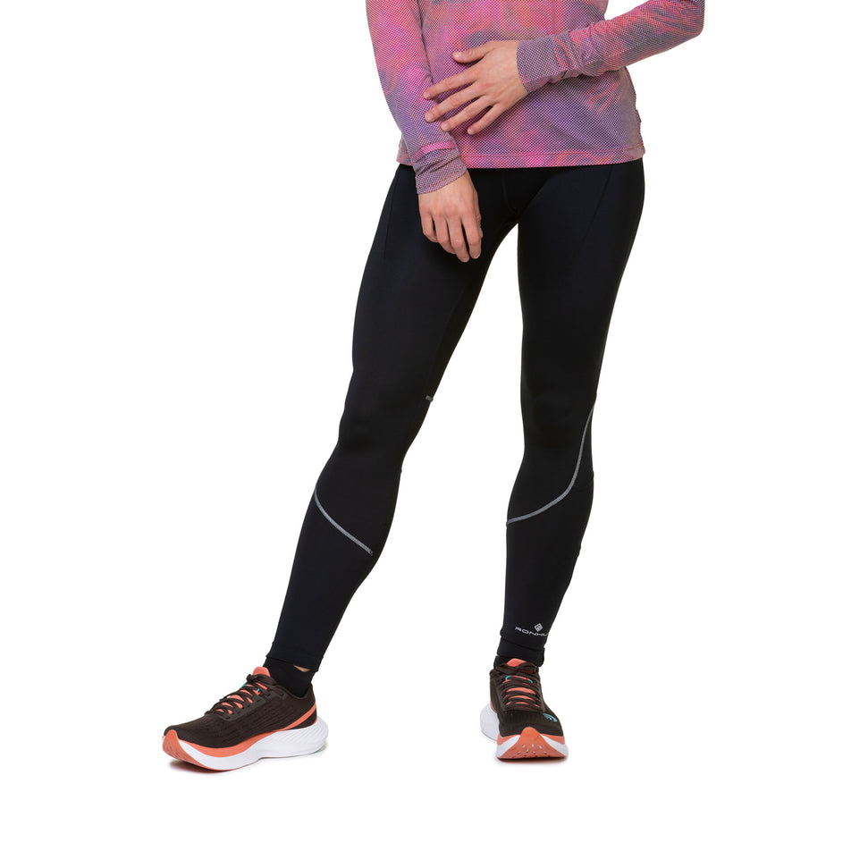 Front view of a model wearing the Ronhill Women's Tech Paragon Tight in the All Black colourway. Model is also wearing shoes and a top. (8489936912546)