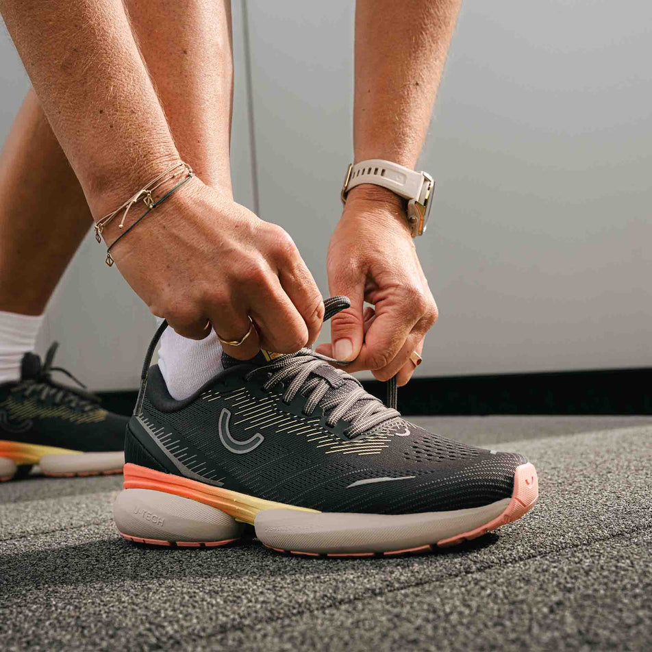 A model tying the laces of the right shoe from a pair of True Motion Women's U-Tech Nevos 3 Running Shoes in the Asphalt/Antarctica/Peach Amber colourway. (8636671459490)