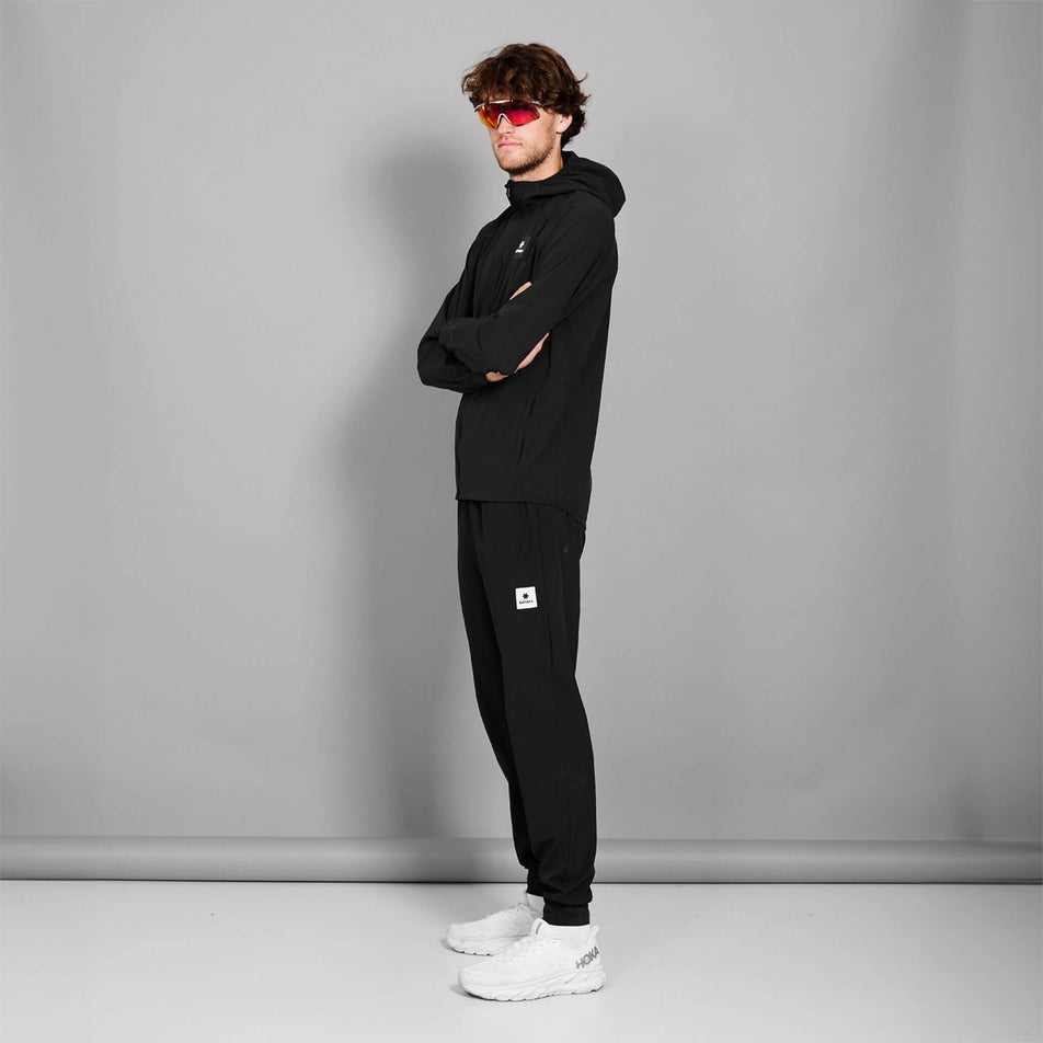 Side view of a model wearing the SAYSKY Men's Clean Pace Flex Hooded Jacket in the Black colourway. Model is also wearing sunglasses, trousers and shoes. (8601312362658)