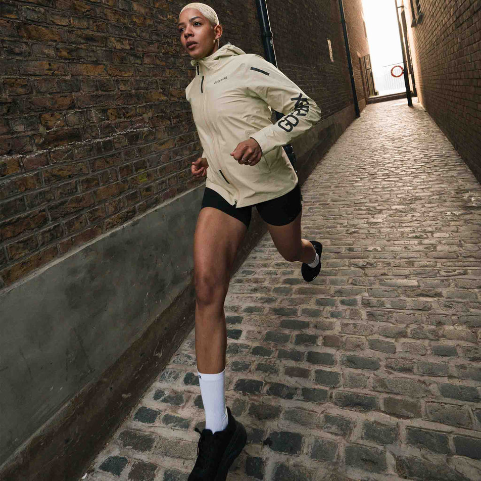 A model running in the GOREWEAR Women's Concurve GORE-TEX Logo Jacket in the Tech Beige/Black colourway. (8752081207458)