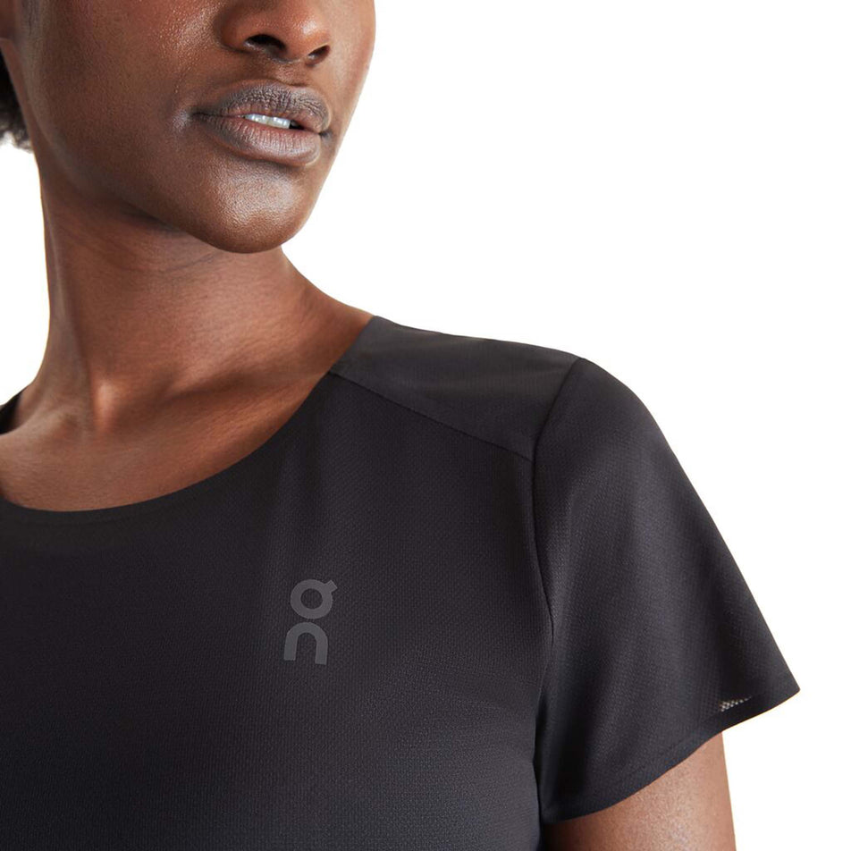 Close-up front view of a model wearing an On Women's Performance T in the Black/Wash colourway. Left side of the upper part of the t-shirt is visible, with the On logo on display.  (8002756313250)