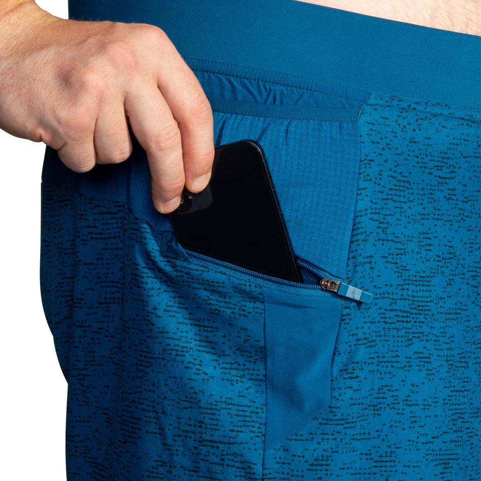 A model demonstrating that, on the right-side of a pair of Brooks Men's Sherpa 7 Inch 2-in-1 Shorts in the Dark Ocean Terrain Print colourway, there is a zipped pocket designed for a phone. (8007477133474)