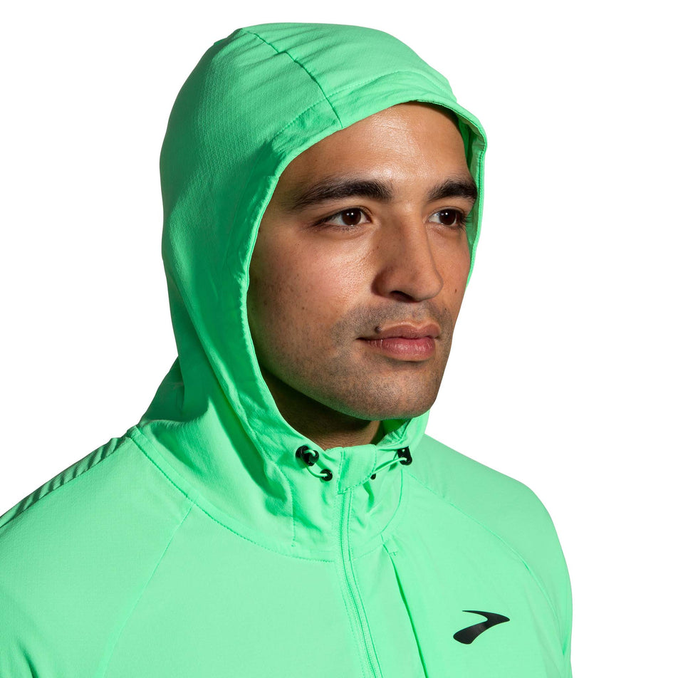 Angled front view of a model wearing a Brooks Canopy Running Jacket in the Neo Green colourway. The model is wearing the jacket with the hood up. Only the top third of the jacket is visible.  (8007418314914)
