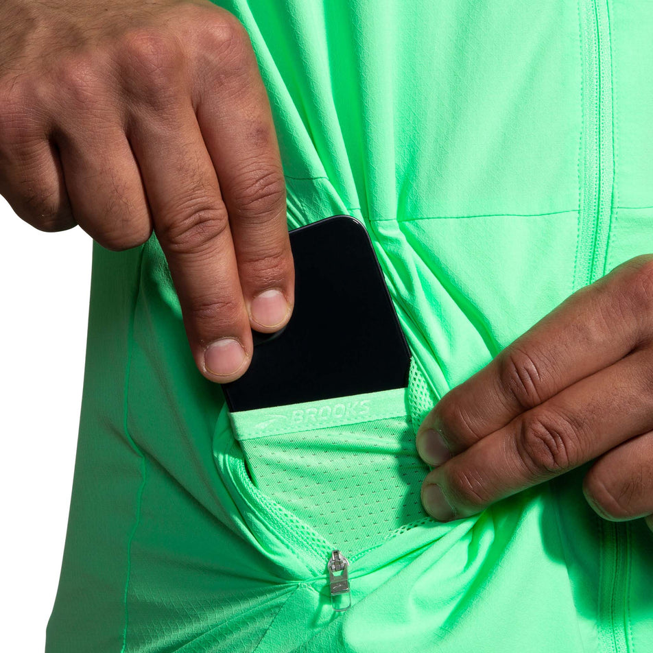 A model demonstrating that there is a pocket for a phone, within the zipped pocket on the lower right side of the front of the Brooks Men's Canopy Jacket in Neo Green (8007418314914)
