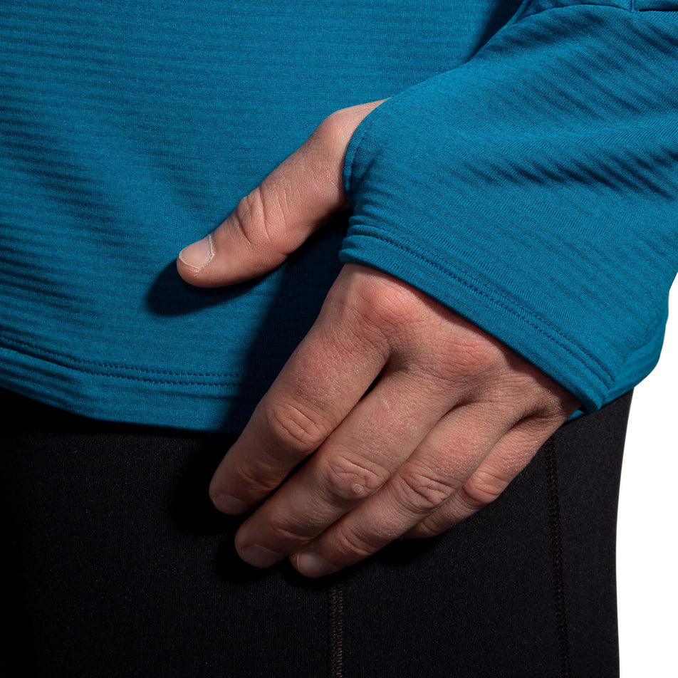 A model wearing a Brooks Men's Notch Thermal Hoodie 2.0 in the Heather Dark Ocean/Dark Ocean colourway, demonstrating that the top has thumb loops at the end of the sleeves.  (8007425589410)