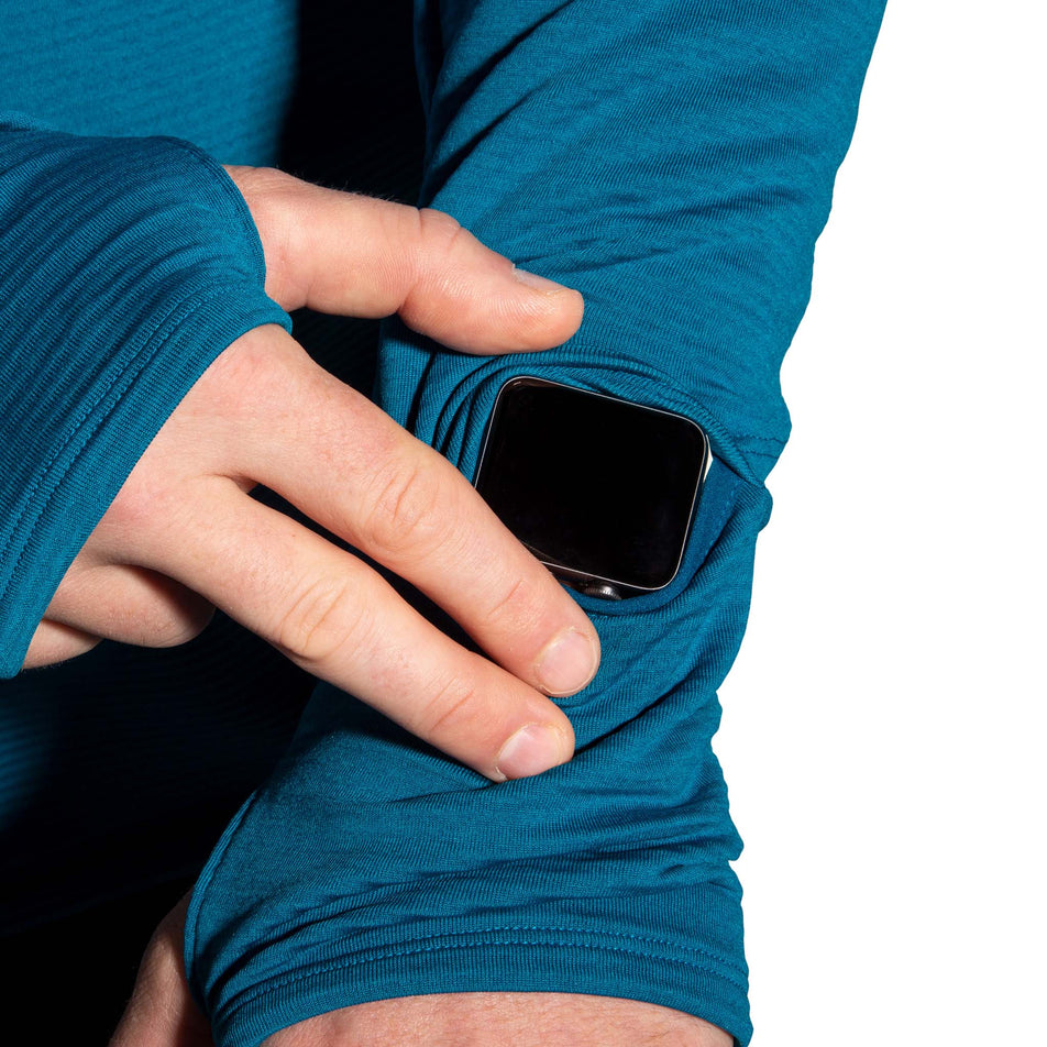 A model wearing a Brooks Men's Notch Thermal Hoodie 2.0 in the Heather Dark Ocean/Dark Ocean colourway, demonstrating that there is a gap in the left sleeve for a running watch to fit through. (8007425589410)