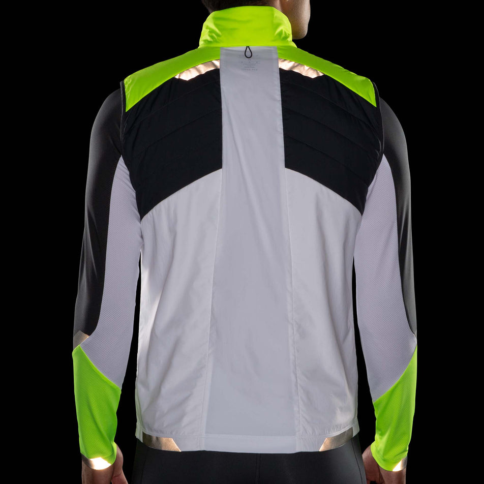 Back view of model wearing a Brooks Men's Run Visible Insulated Vest in the White/Asphalt/Nightlife colourway. Model is wearing also wearing a Brooks long sleeve top. Items being worn in dark conditions to show the reflectivity. (8059815329954)