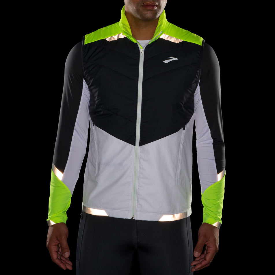 Front view of model wearing a Brooks Men's Run Visible Insulated Vest in the White/Asphalt/Nightlife colourway. Model is wearing also wearing a Brooks long sleeve top. Items being worn in dark conditions to show the reflectivity. (8059815329954)
