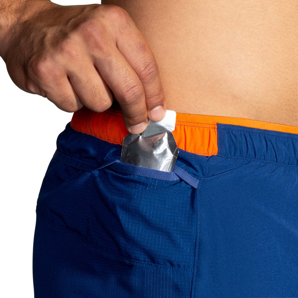 A model demonstrating that an energy gel can be stored in one of the mesh side pockets on the right-hip section of a pair of Brooks Men's High Point 7