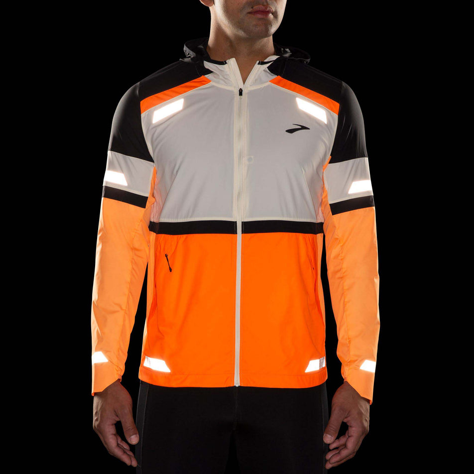 Front view of a model wearing 
the Brooks Men's Run Visible Jacket 2.0 in the Ecru/Fluoro Flash/Black colourway. Image shows what reflective strips look like when artificial light hits them. (8499396804770)