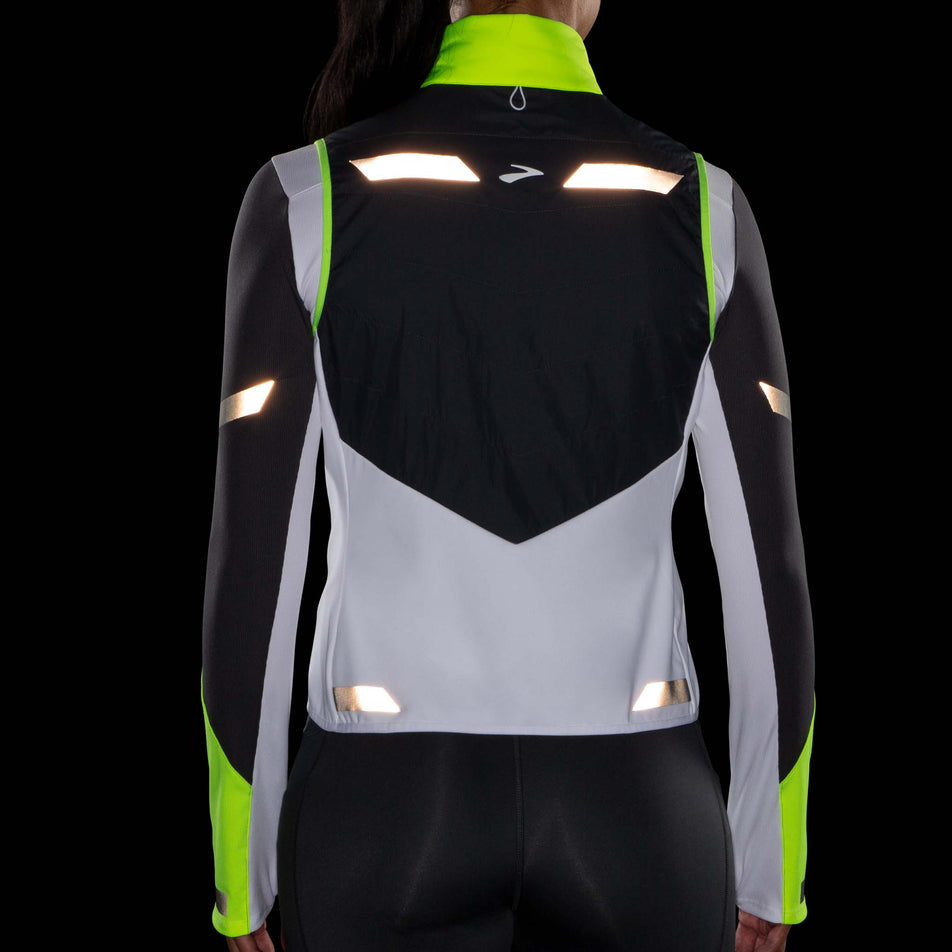 Back view of a model wearing a Women's Run Visible Insulated Vest in the White/Asphalt/Nightlife colourway. Model is also wearing a Brooks long sleeve top. The clothing is being worn in dark conditions to show the clothing's reflectivity.  (8059825914018)