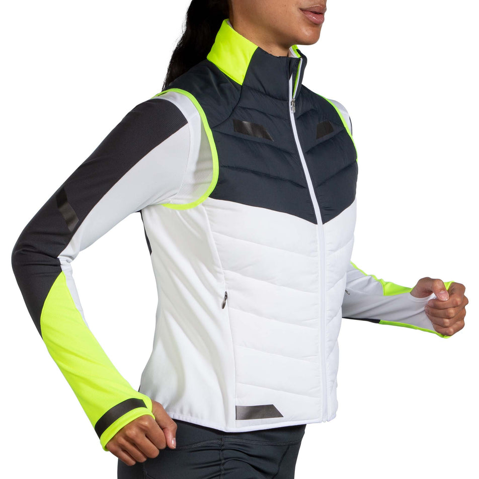 Side view of a model wearing a Women's Run Visible Insulated Vest in the White/Asphalt/Nightlife colourway. Model is also wearing a Brooks long sleeve top. (8059825914018)