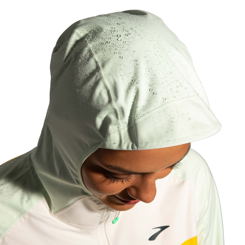 Drops of water on the hood of a Brooks Women's High Point Waterproof Jacket in the Lemon/Ecru/Glacier Green colourway. The jacket is being worn by a model. (8177453367458)