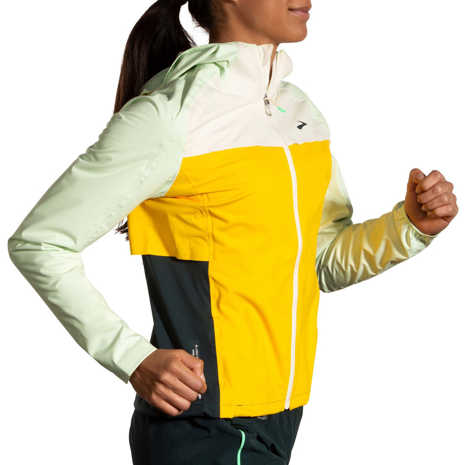 Side view of a model wearing a Brooks Women's High Point Waterproof Jacket in the Lemon/Ecru/Glacier Green colourway. Model is also wearing Brooks legwear. (8177453367458)